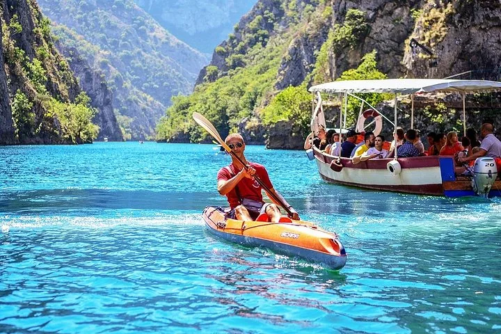 A kayaker paddles on the bright blue waters of Matka Canyon alongside a tour boat.
